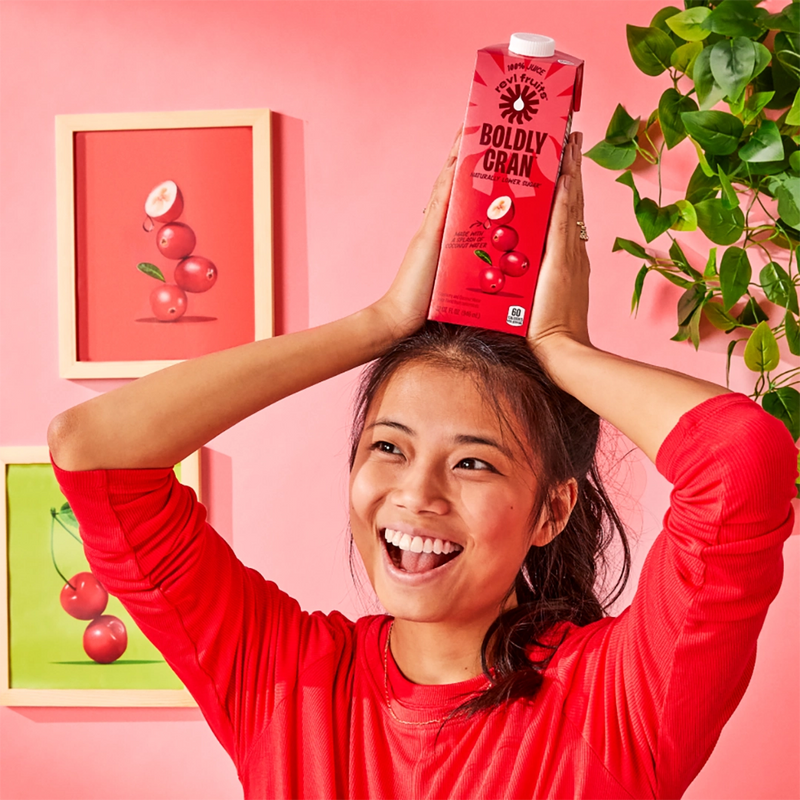 Smiling woman holding a carton of Revl Fruits juice on her head against a pink background.